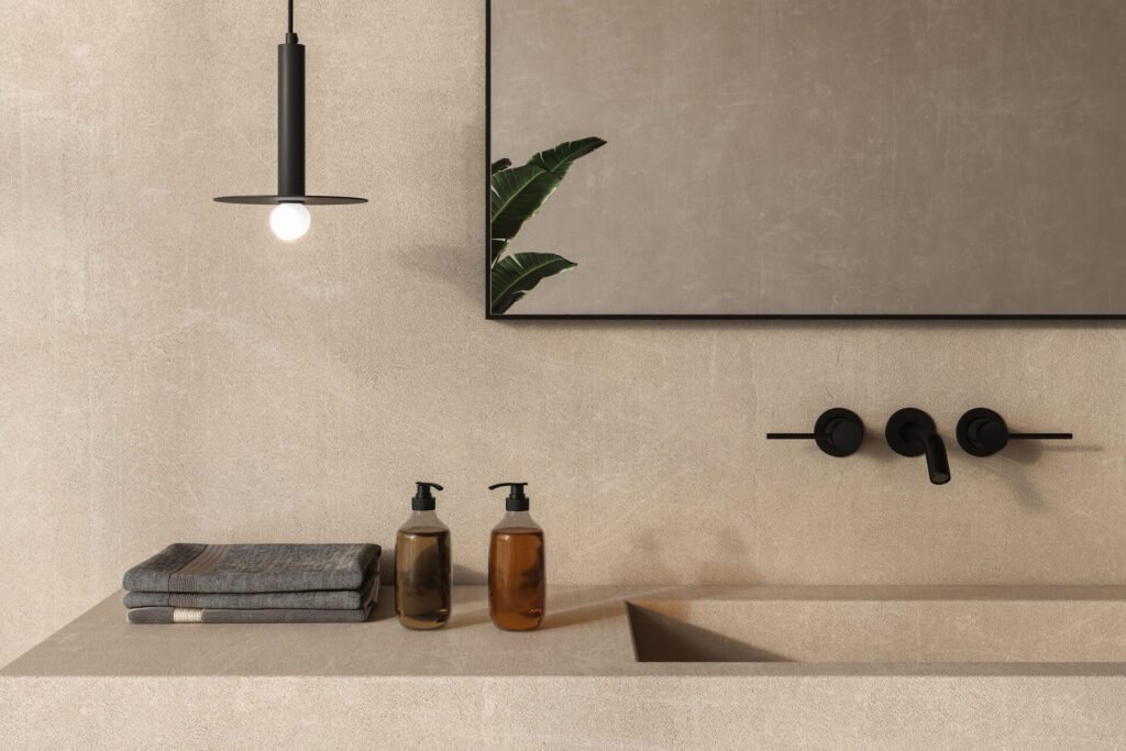 Close-up of an earthy bathroom vanity with beige stone wall and countertop, black pendant light, soap containers, folded hand towels, and a plant reflected in the mirror.