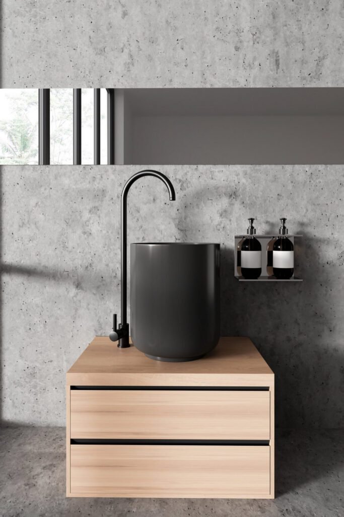 Portrait image of a minimalist bathroom featuring a light wood vanity with a dark grey sink and tall faucet against a grey stone wall and floor.