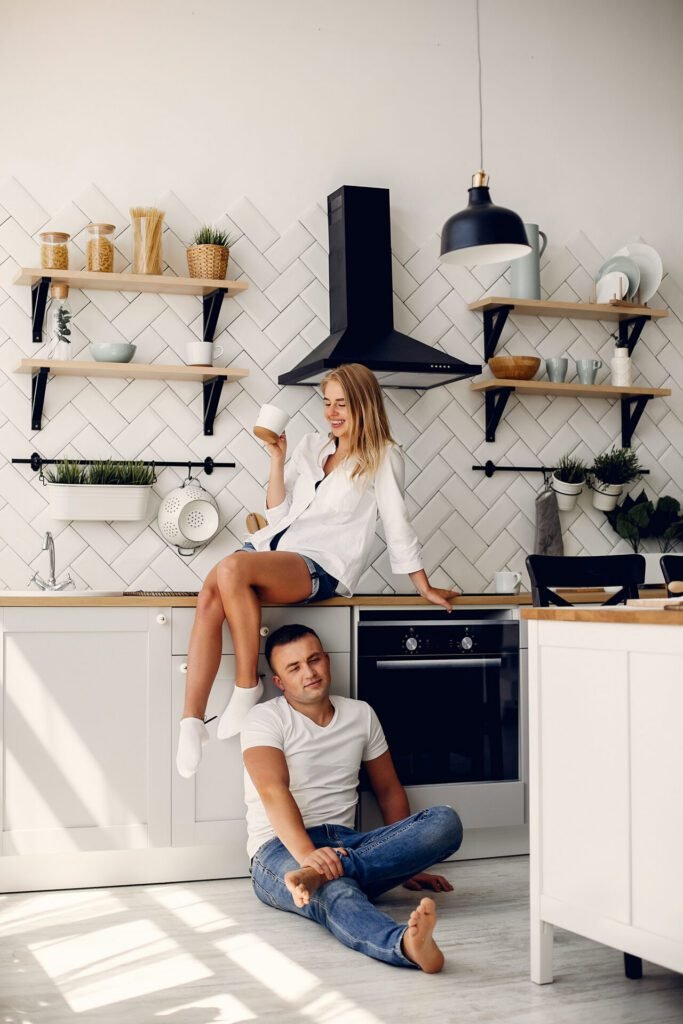 Portrait of a woman sitting on the counter with a coffee cup and a man sitting on the floor in their rustic modern kitchen with white cabinets, wood countertops, and black accents.