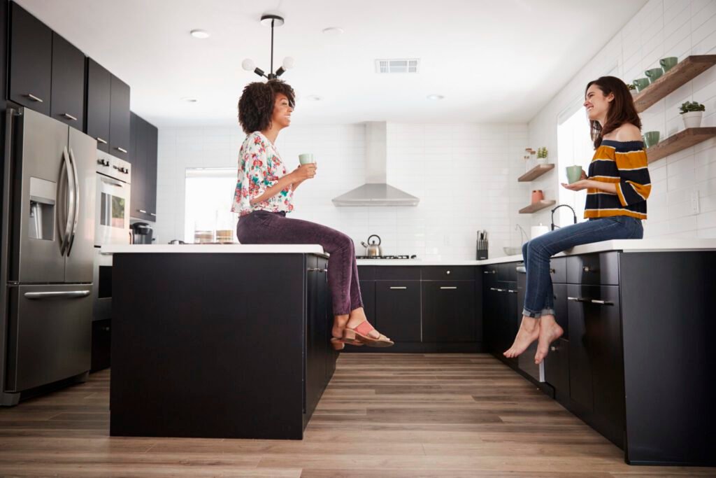 Two female friends sitting on kitchen countertops, smiling and sipping hot drinks while having a friendly conversation.