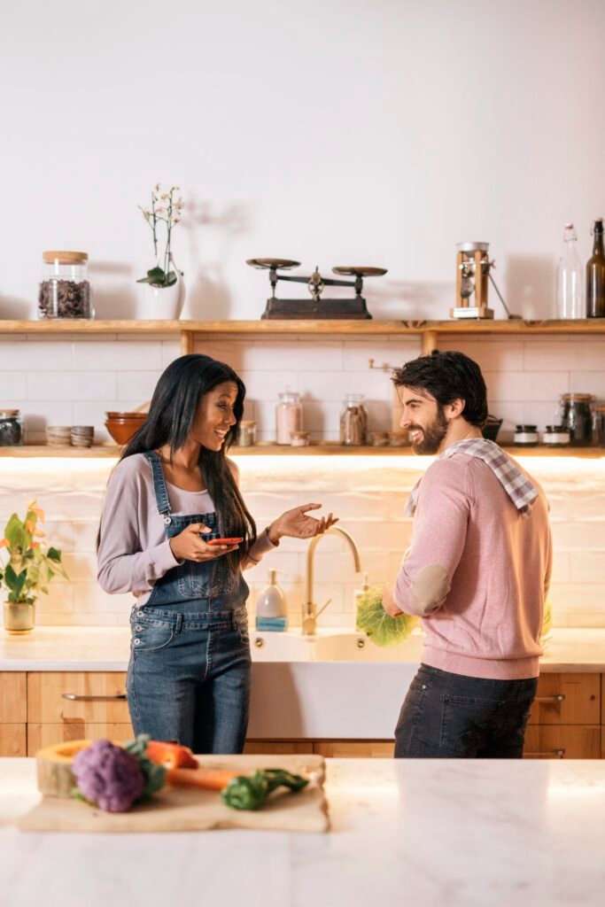 Portrait image of a smiling couple talking in their new kitchen with a white farm sink, white countertops, and vegetables on a cutting board on the island.