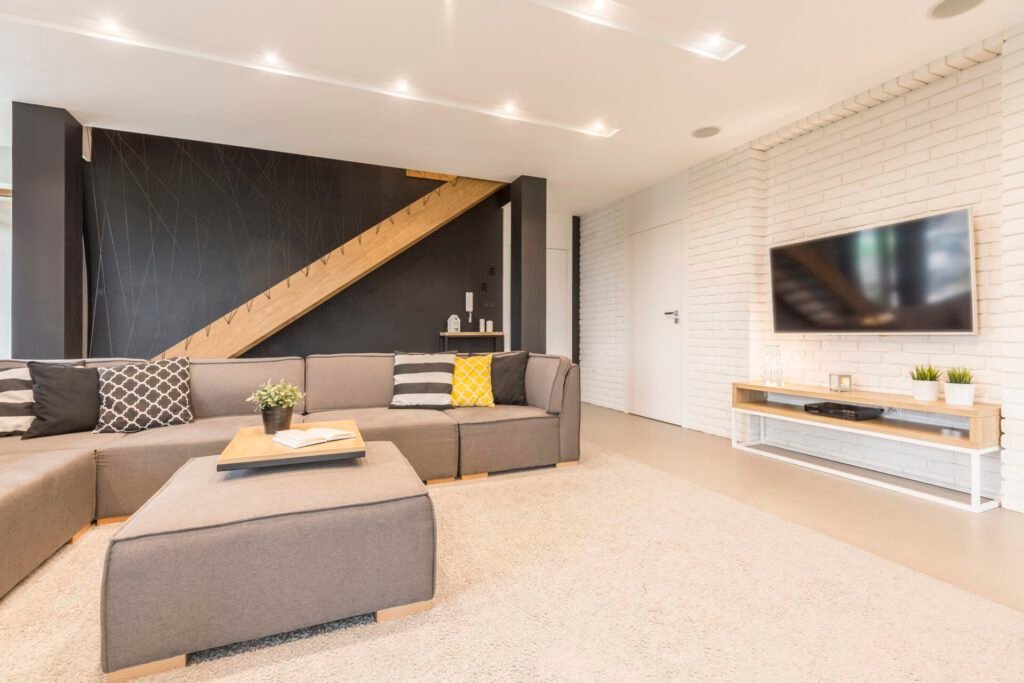 Modern finished basement with a large flat-screen TV, sectional couch, and stairs visible in the background.